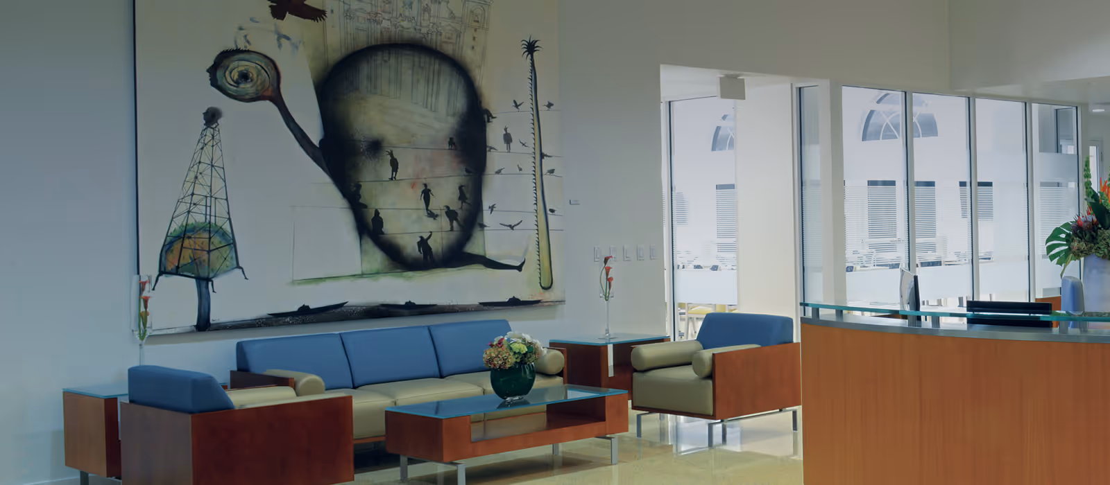 A modern reception lounge with blue and beige seating, a glass-topped wooden coffee table, large abstract wall art, and a curved wooden reception desk by floor-to-ceiling windows.