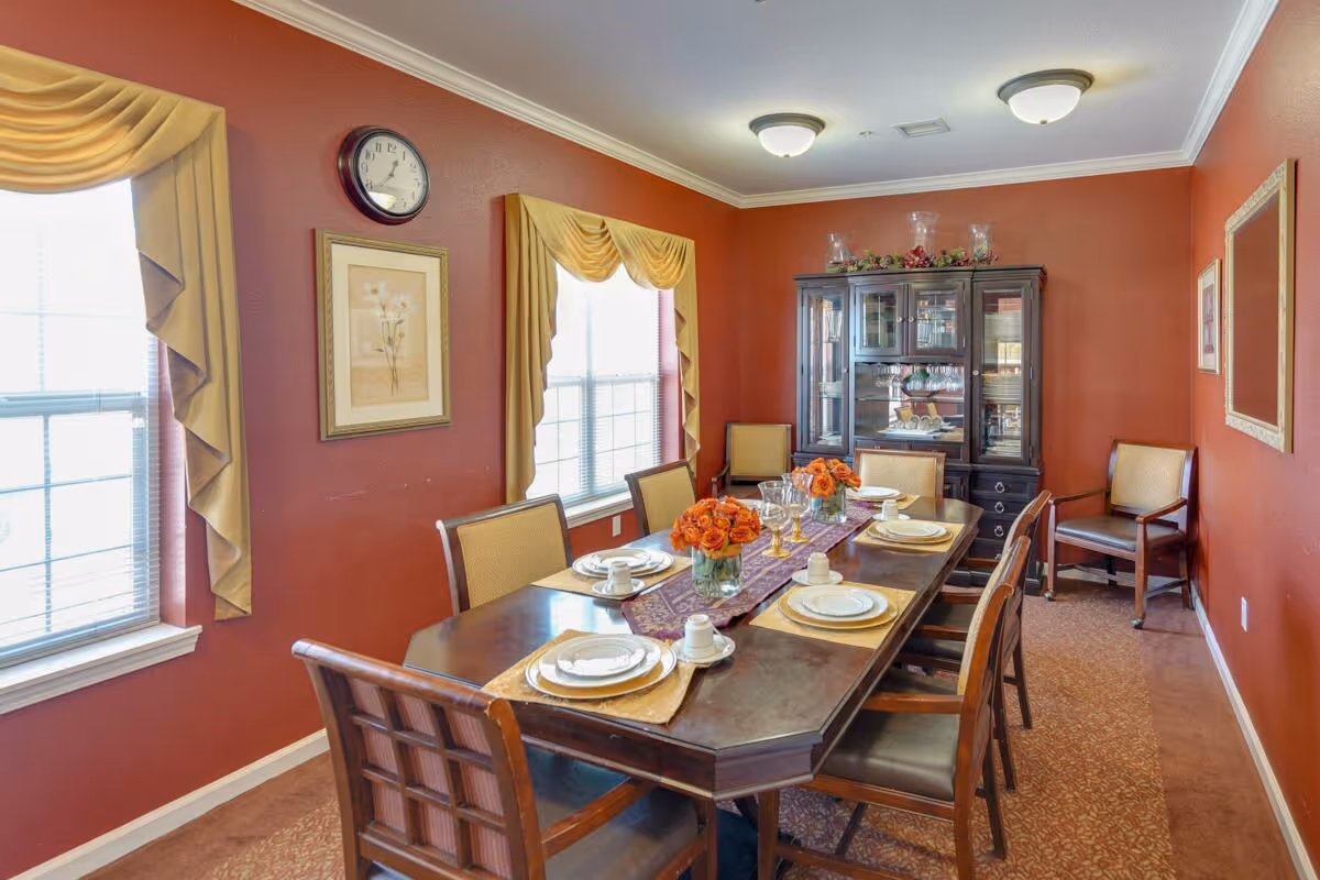 A dining room with a long wooden table set for six people, featuring white plates, cups, and gold-rimmed glasses. The table has a purple and gold runner with two vases of orange flowers. The room has red walls, two windows with yellow drapes, a wall clock, framed artwork, and a dark wooden china cabinet filled with dishes and glassware. There are six chairs around the table and one additional chair against the wall.