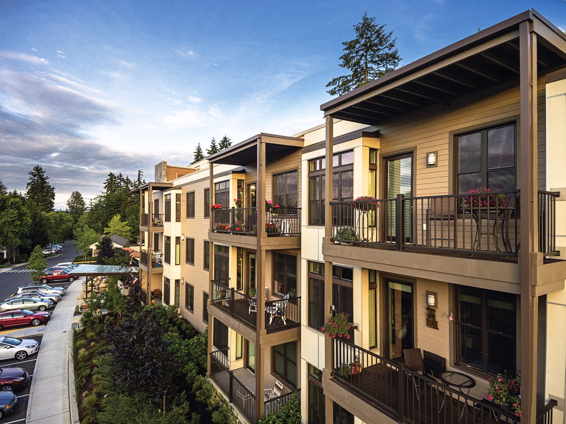 Exterior view of a multi-story residential building with balconies adorned with potted plants and outdoor furniture, surrounded by trees and a parking lot with cars under a partly cloudy sky.