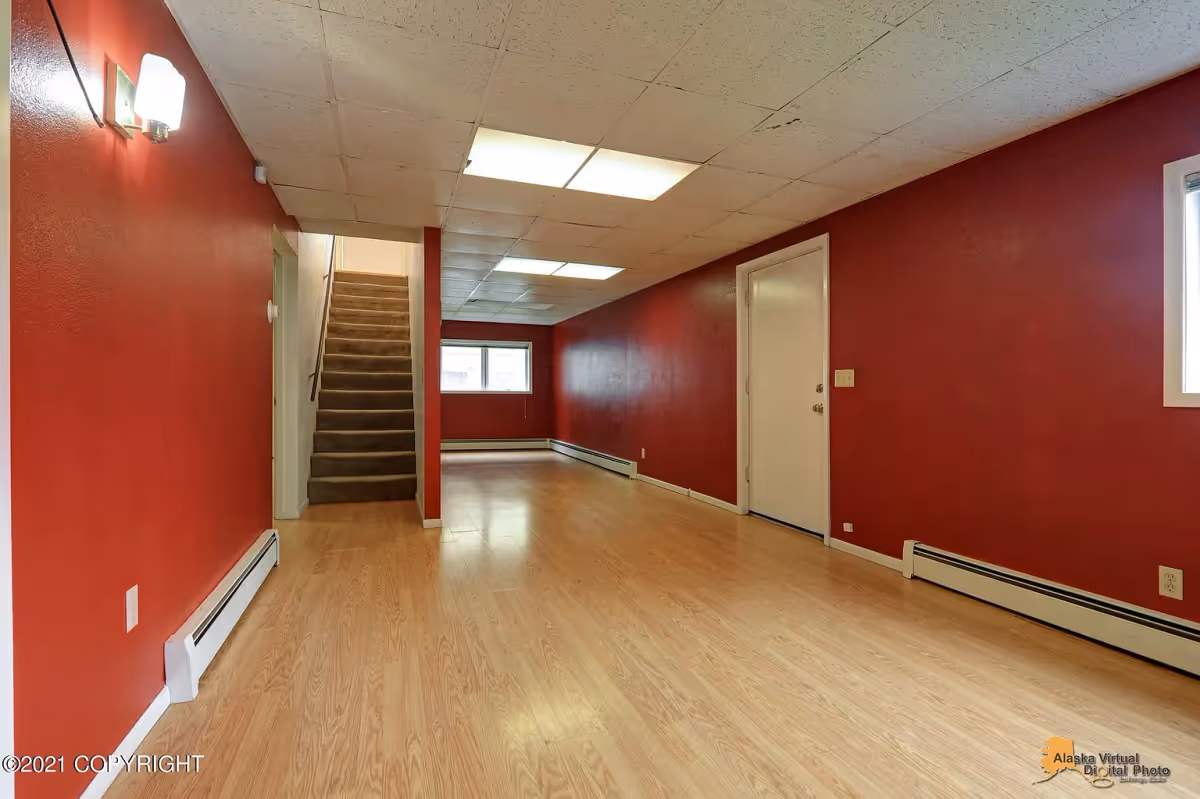 Empty interior room with red walls, light wood flooring, and a white door on the right. There is a staircase with carpet leading upwards at the back left, fluorescent ceiling lights, and a window at the far end of the room.