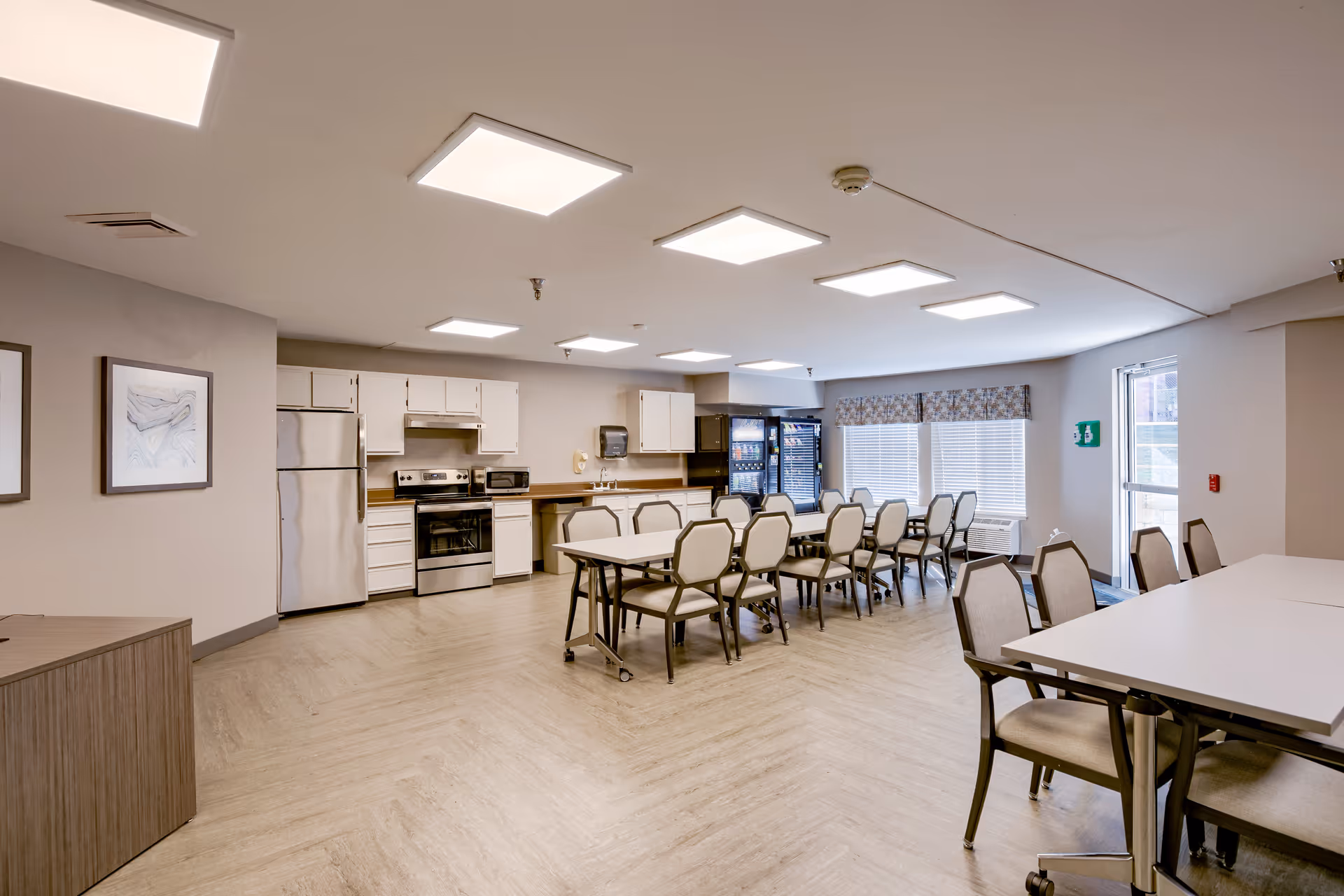 A bright communal dining and kitchen area with multiple tables and chairs arranged for group seating. The kitchen features white cabinets, a stainless steel refrigerator, stove, and microwave. Large windows with blinds and a patterned valance allow natural light into the room. The floor has a light wood pattern, and the ceiling has multiple square recessed lights.