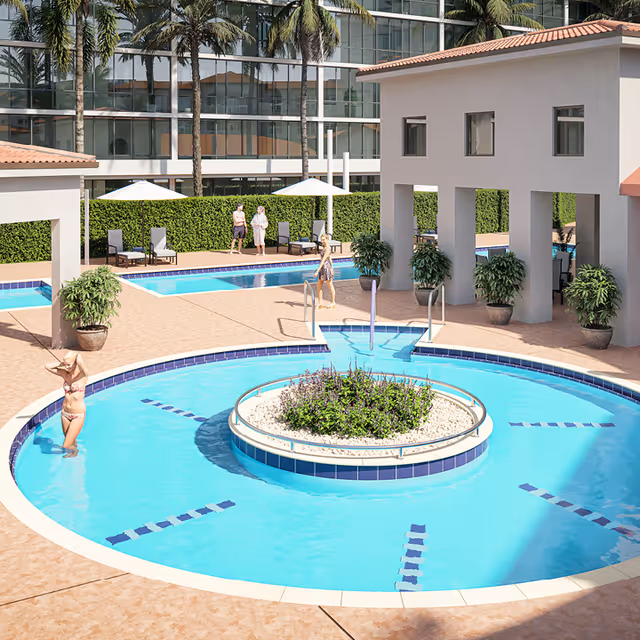 Outdoor swimming pool area with a circular pool featuring a central planter with greenery. Several people are enjoying the pool and surrounding deck, which has lounge chairs and umbrellas. Palm trees and a modern building with large windows are visible in the background.