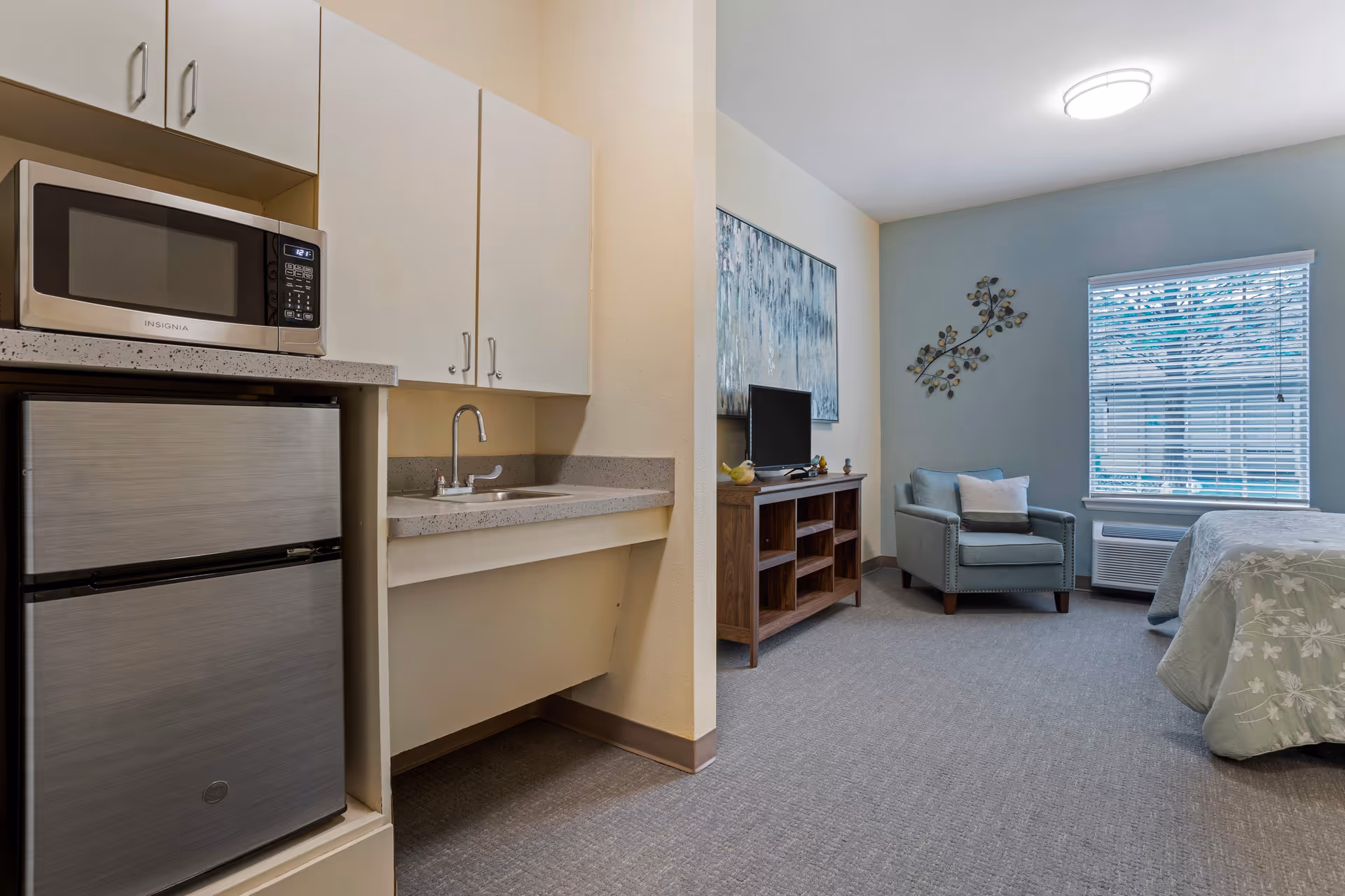 Interior view of a senior living facility room showing a small kitchenette area with a microwave and mini refrigerator on the left, and a living space with a blue armchair, a wooden TV stand with a television, a large window with blinds, and part of a bed with a floral bedspread on the right.
