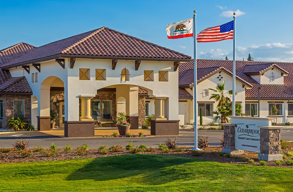 Exterior view of Cedarbrook Memory Care Community building with a tiled roof, beige walls, and a covered entrance supported by columns. Two flagpoles display the California state flag and the United States flag. There is a sign in front of the building with the facility name, surrounded by landscaped grass and plants under a clear blue sky.