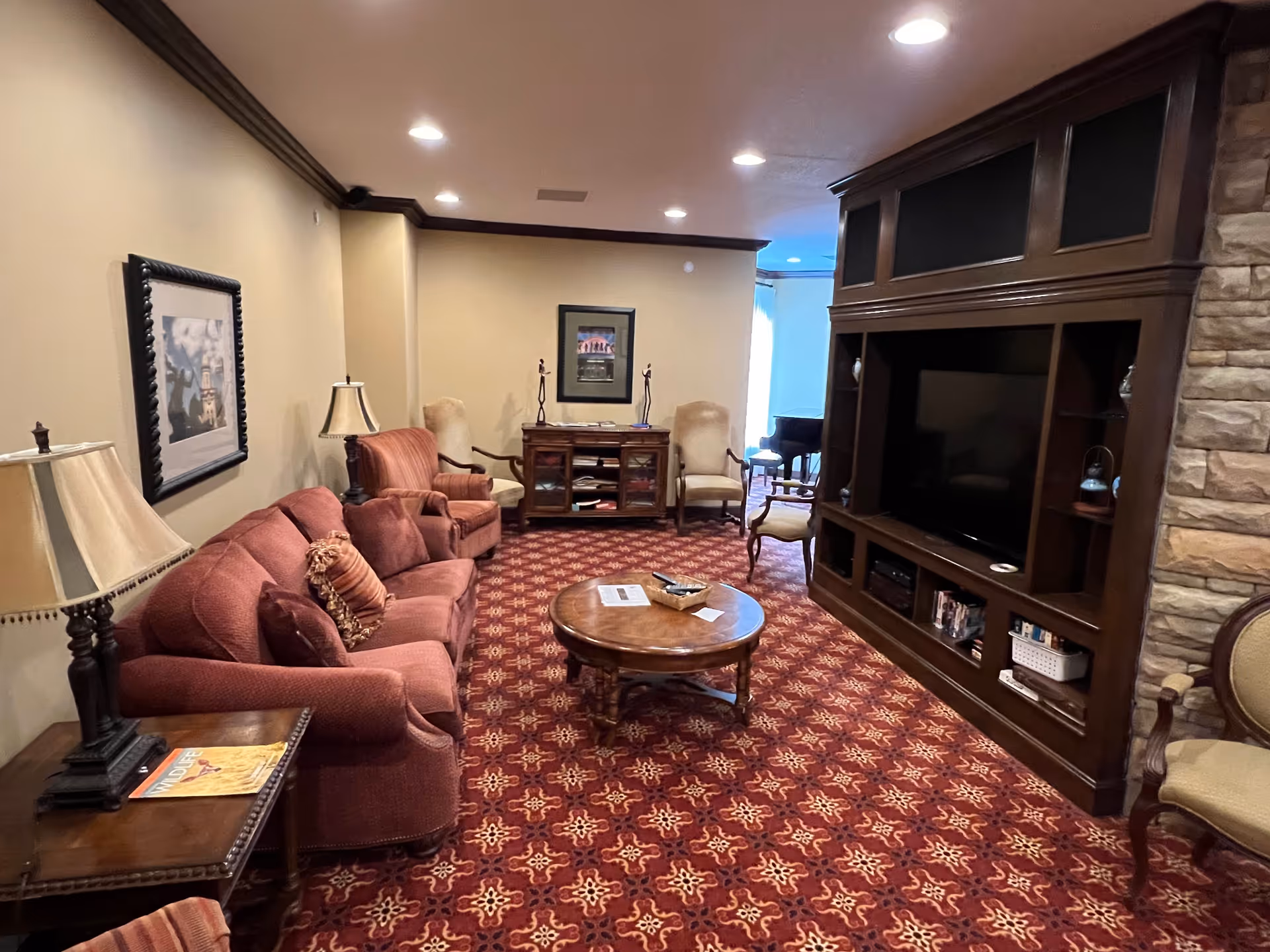 A cozy senior living community lounge with red patterned carpet, a red sofa, armchairs, a wooden coffee table, and a large dark wood entertainment center with a TV. The room is softly lit with recessed ceiling lights and decorated with framed artwork and table lamps.