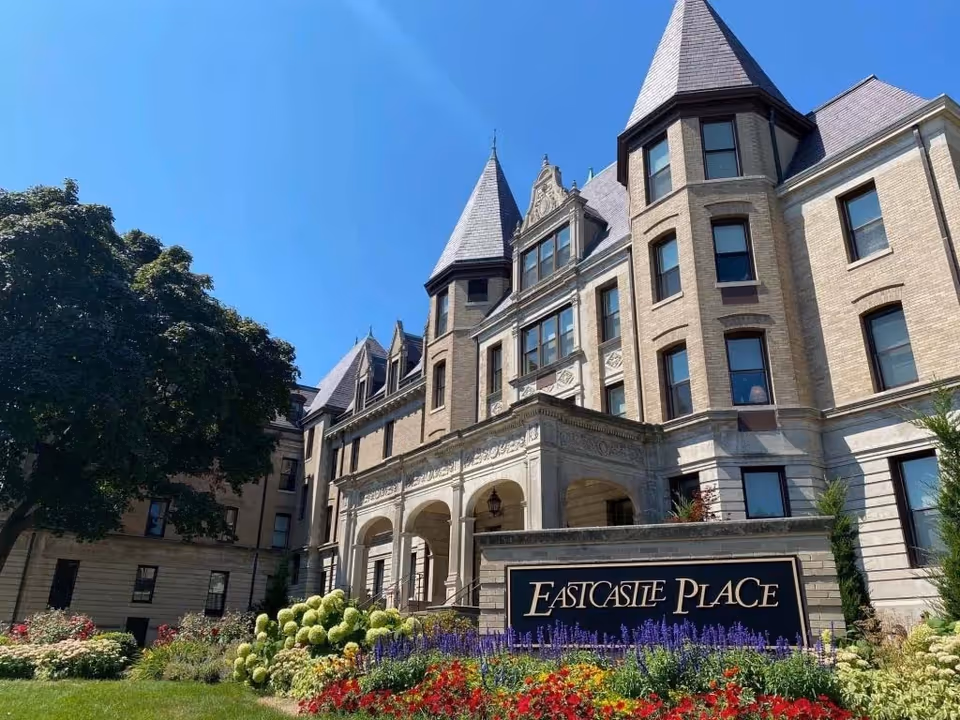 Historic multi-story brick building with turrets, landscaped flowerbeds, and a front sign reading 'Eastcastle Place'.
