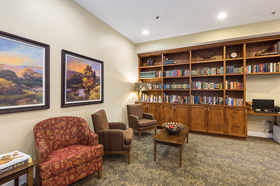 A cozy library or reading room with a large wooden bookshelf filled with books, two framed landscape paintings on the wall, three upholstered chairs with floral and striped patterns, a wooden coffee table with a decorative bowl, and a small side table with a book titled 'Songs Bible'.