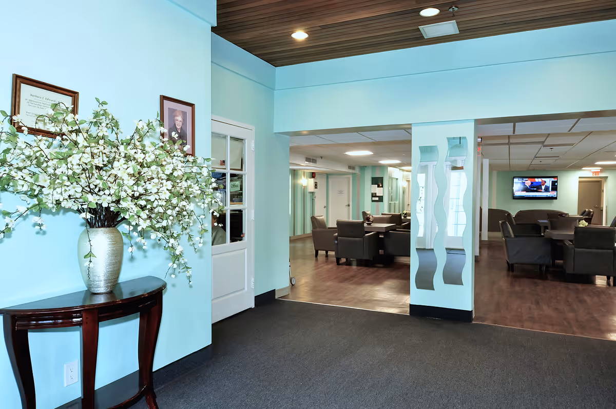 Interior view of a senior living facility lobby area with light blue walls, a wooden table holding a large vase of white flowers, framed pictures on the wall, and an open doorway leading to a lounge area with several dark armchairs and a wall-mounted TV.