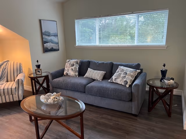 A cozy living room with a gray sofa adorned with three decorative pillows, a glass-top coffee table with decorative items, two round wooden side tables each holding a small lamp and decor, a striped armchair, a large window with blinds, and a framed abstract painting on the wall.