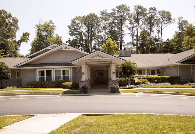 Front exterior view of a single-story building with a covered entrance, surrounded by well-maintained landscaping and tall trees in the background under a clear sky.