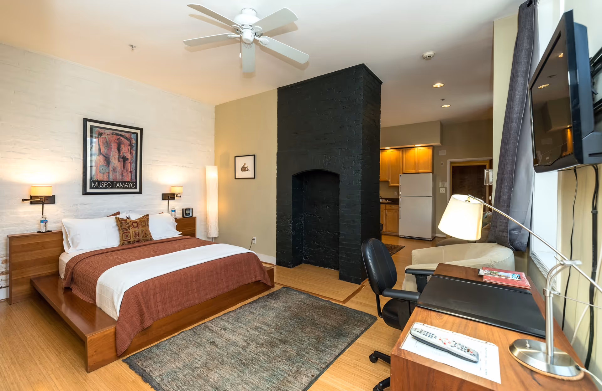 A cozy bedroom featuring a large wooden platform bed with white and brown bedding, two bedside tables with lamps, a black painted brick fireplace in the center, a desk with a lamp and chair, a wall-mounted TV, and a small kitchen area with wooden cabinets and a refrigerator in the background.