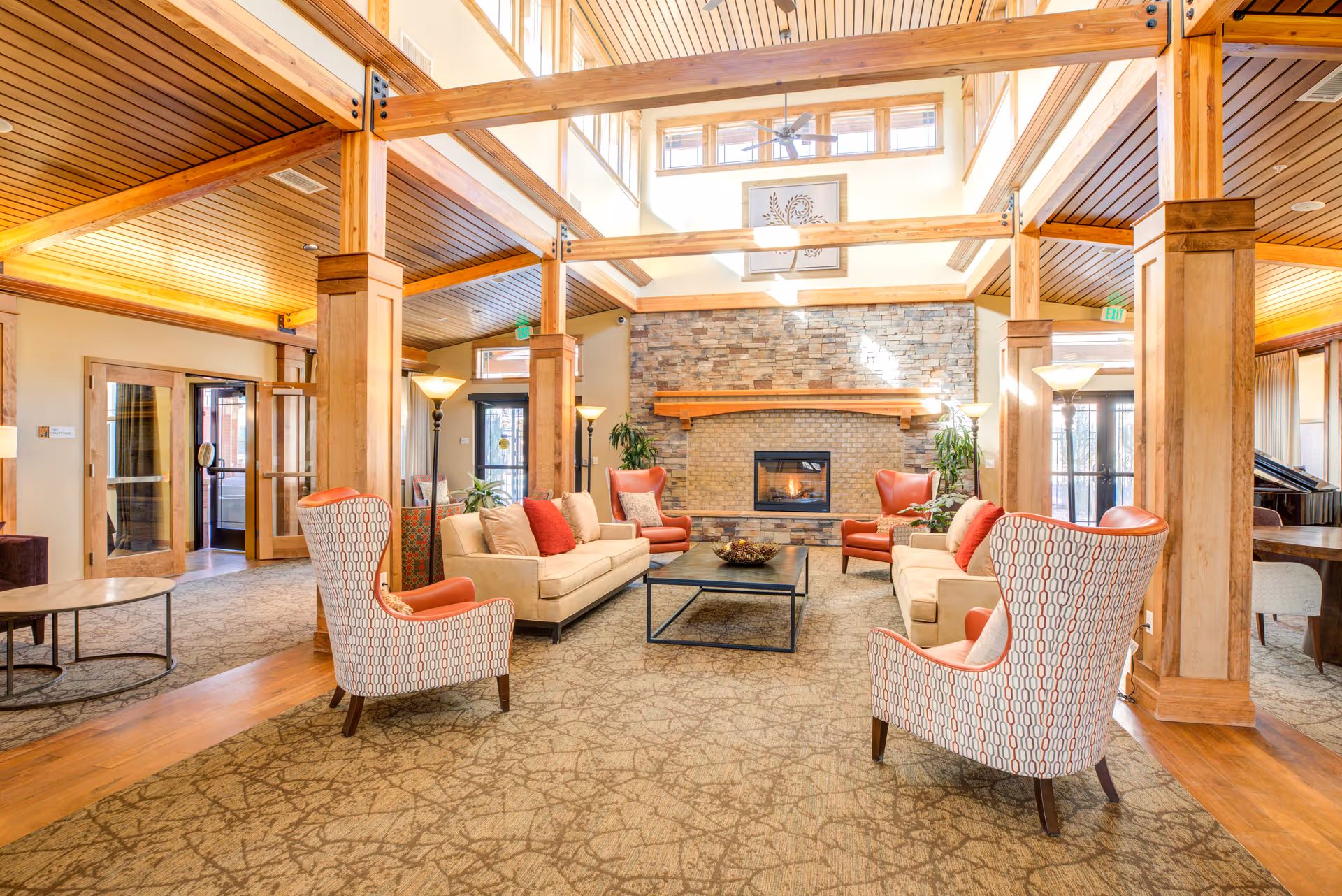 A spacious and well-lit living room area with high ceilings and wooden beams. The room features a stone fireplace with a fire burning, two beige sofas with red and beige cushions, four patterned armchairs with orange cushions, a black coffee table, floor lamps, and large windows letting in natural light. The room has a carpeted floor with a patterned design and wooden accents throughout.