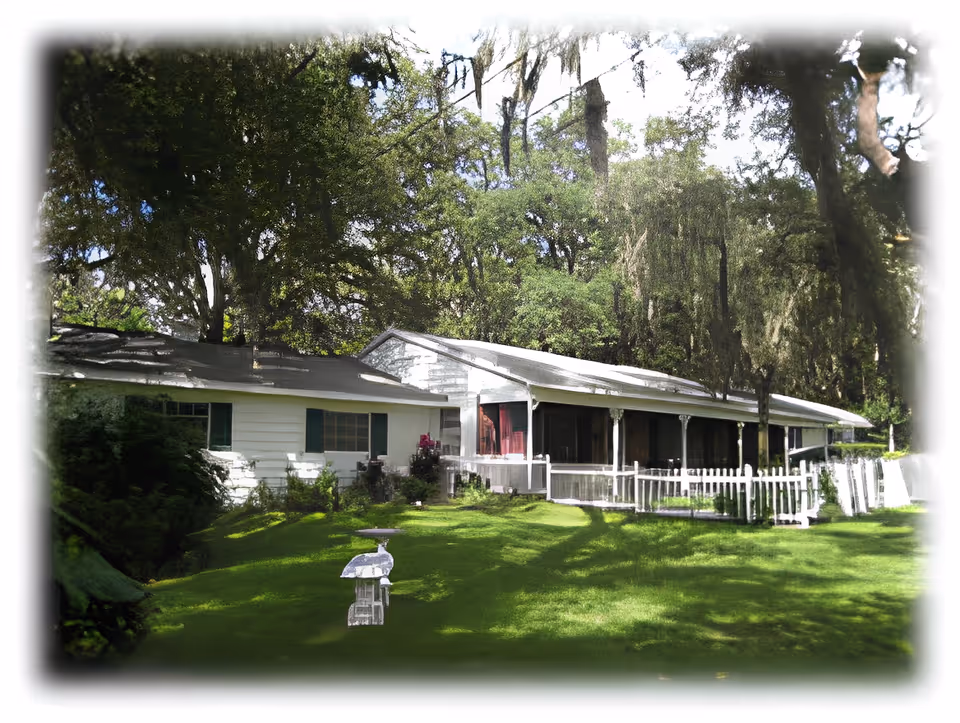 A white single-story building with a long porch surrounded by green grass and large trees with hanging moss, under a partly cloudy sky.