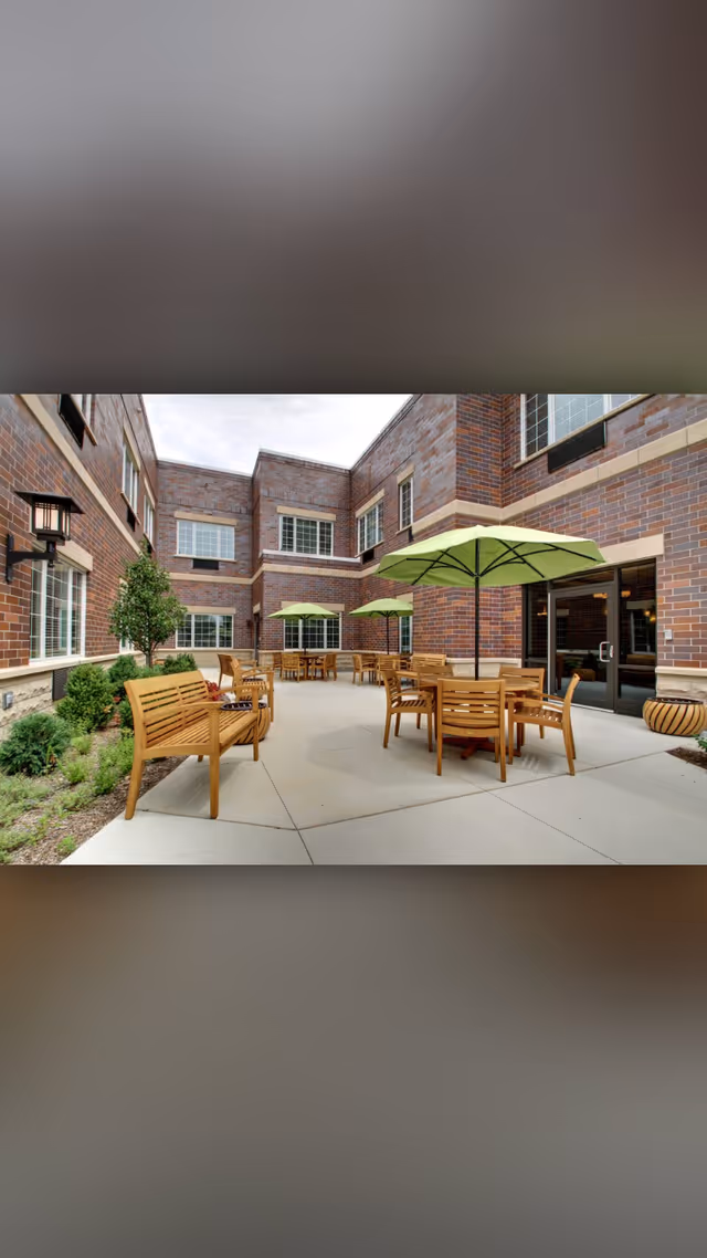 Outdoor courtyard area of a senior living facility with wooden benches and tables, green umbrellas providing shade, surrounded by brick building walls and windows, with some greenery along the edges.