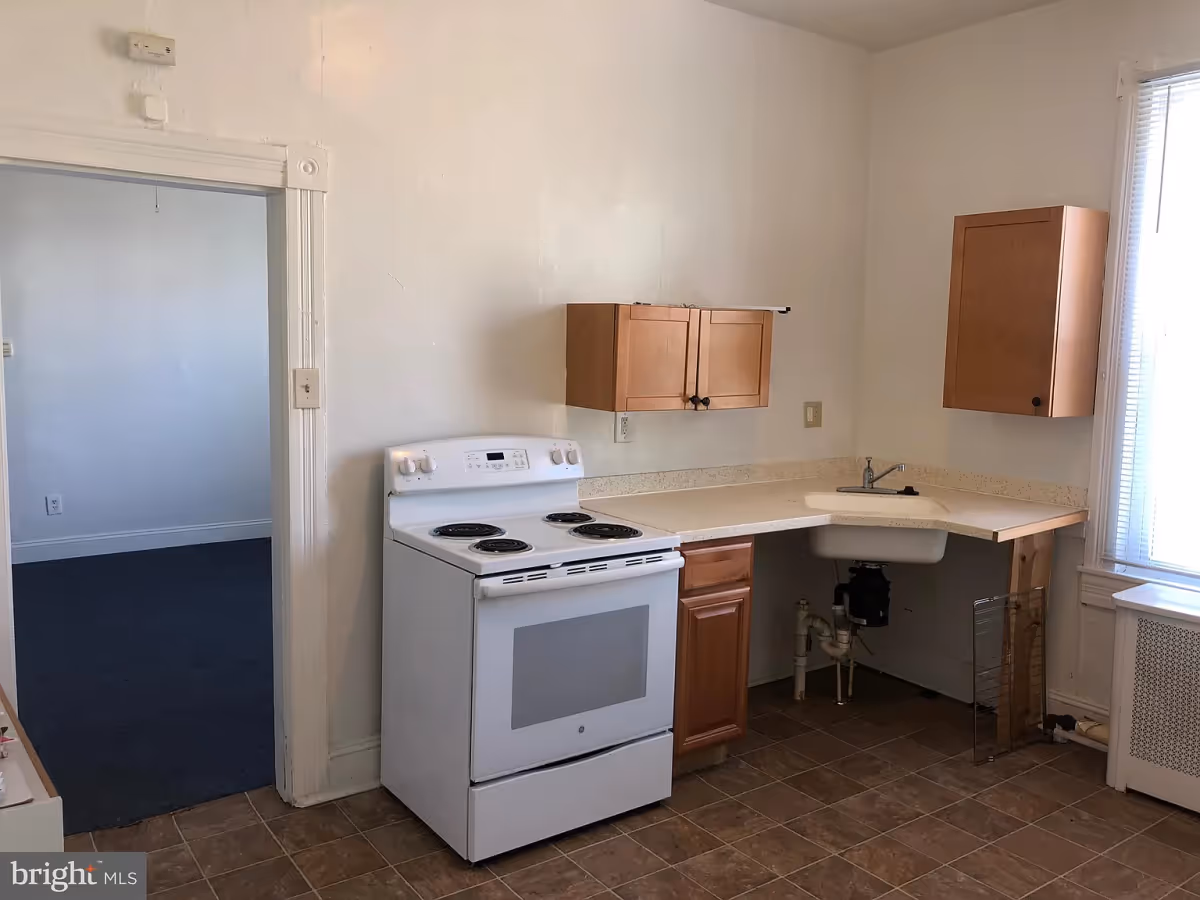 A small kitchen area with a white electric stove, wooden cabinets mounted on the wall and under the counter, a corner sink, and a window with blinds letting in natural light. The floor is tiled, and there is an open doorway leading to another room with carpeted flooring.