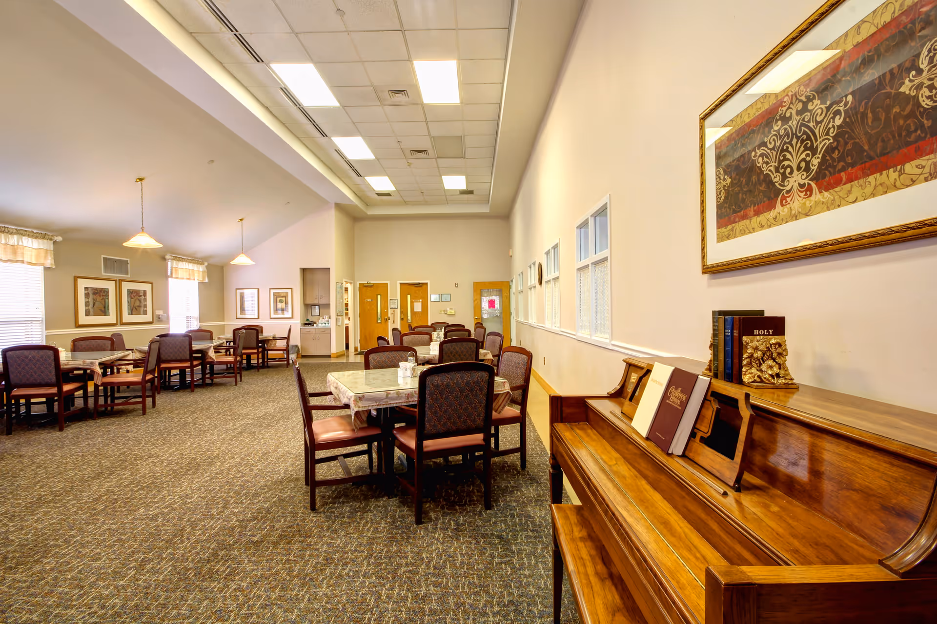 A spacious dining room with multiple tables covered with tablecloths and surrounded by chairs. On the right side, there is a wooden piano with several books on top, including a Holy Bible. The room has carpeted floors, framed artwork on the walls, and large windows with curtains allowing natural light to enter. The ceiling features recessed lighting and hanging pendant lights.