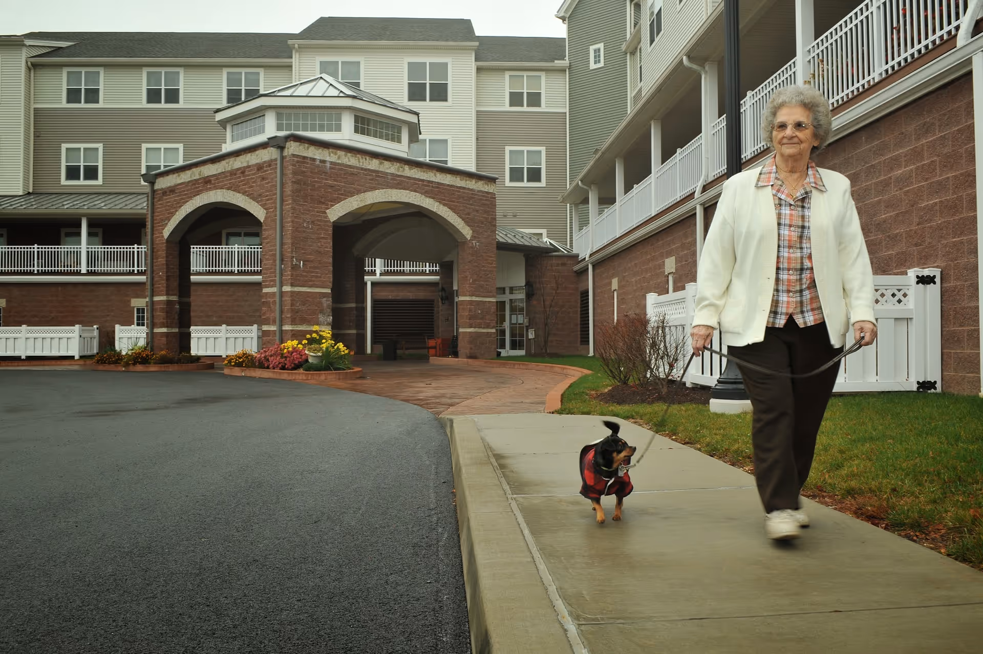 An elderly woman wearing a white jacket and plaid shirt walking a small dog on a leash along a sidewalk outside a multi-story senior living facility with brick and siding exterior.