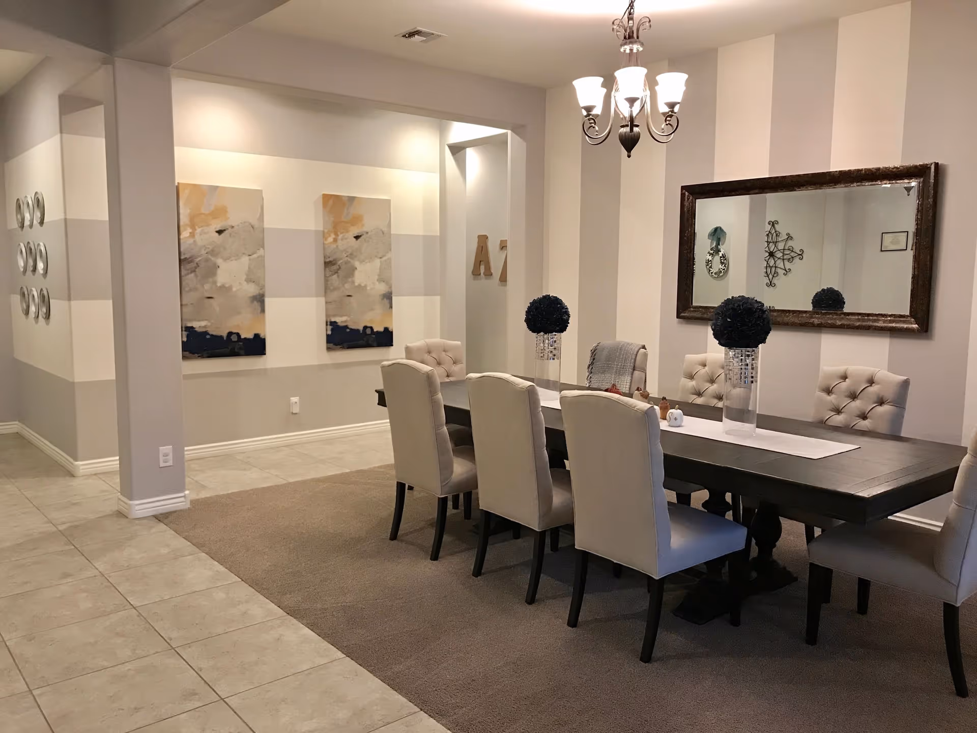 A dining room with a long dark wooden table surrounded by eight beige upholstered chairs. The room features a striped wall with a large rectangular mirror and a chandelier hanging above the table. Two decorative vases with dark spherical floral arrangements are placed on the table. The floor is a combination of tile and carpet, and there are two abstract paintings on the adjacent wall.