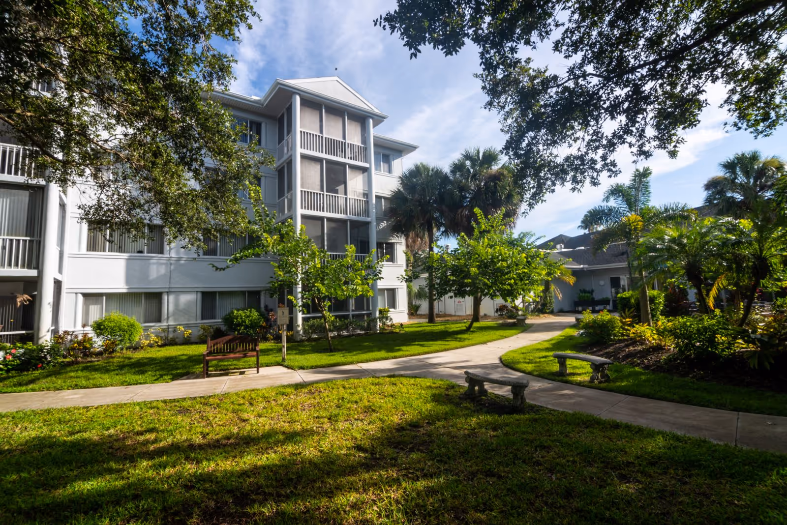 Exterior view of a senior living facility with a white multi-story building surrounded by green trees, bushes, and a well-maintained lawn. There are paved walkways, benches, and a clear blue sky with some clouds.