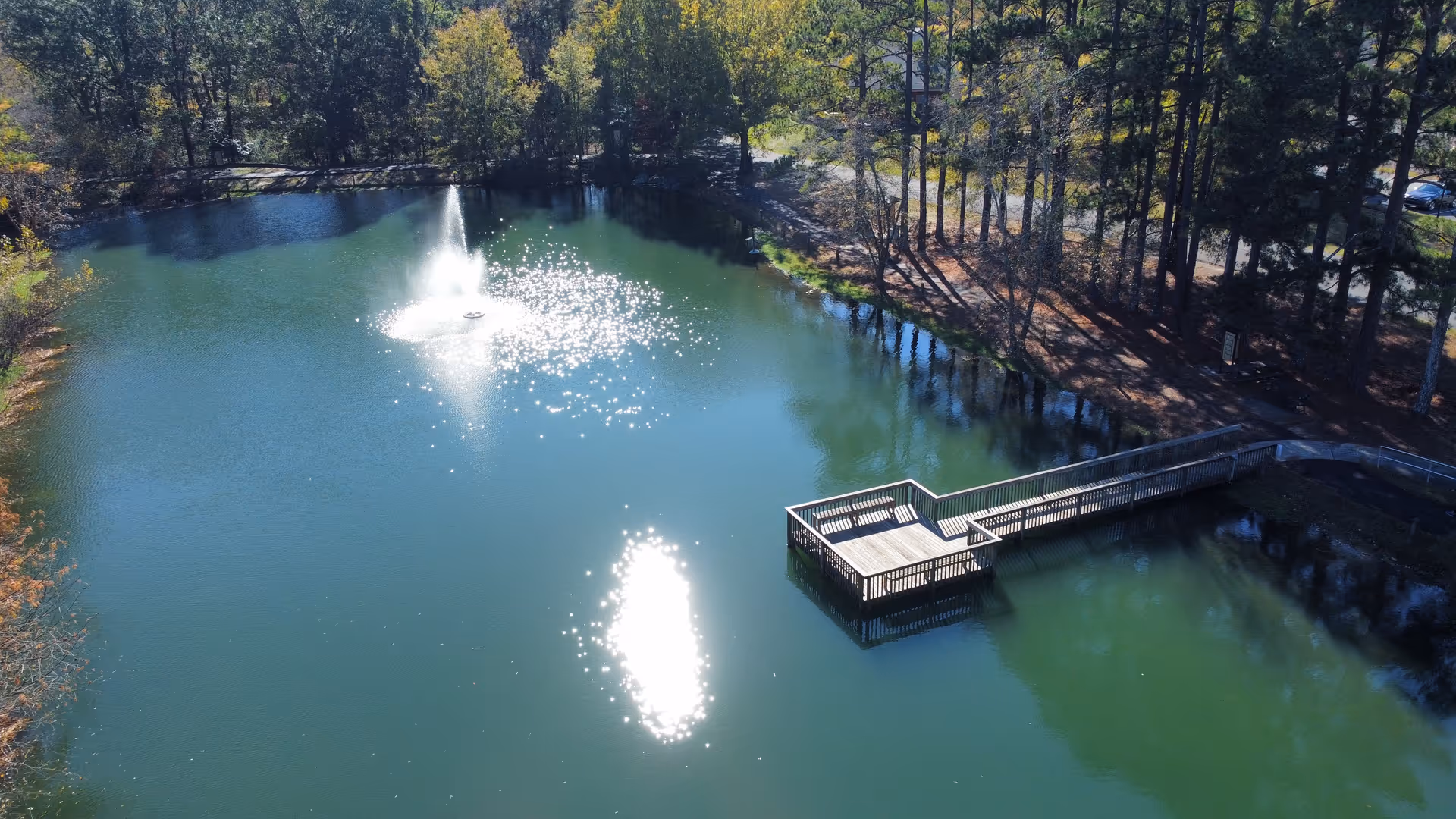 A serene pond surrounded by trees with a wooden dock extending over the water and a fountain spraying water in the center, reflecting sunlight.