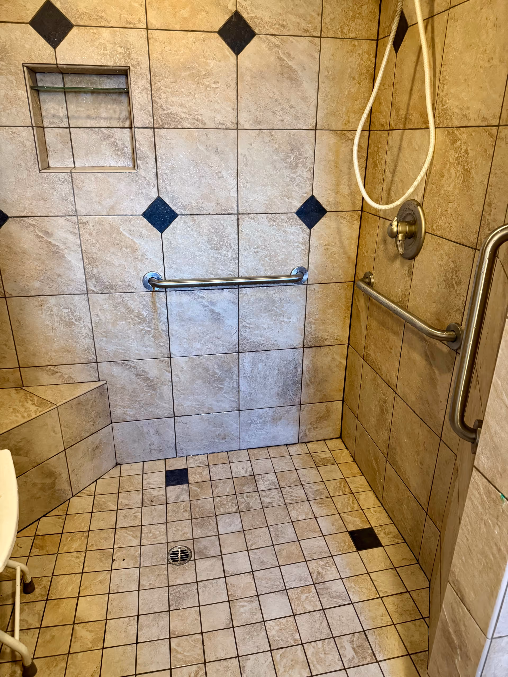 A tiled walk-in shower with beige and black square tiles on the walls and floor. The shower features two metal grab bars, a built-in tiled bench, a recessed shelf, a handheld showerhead with a white hose, and a drain in the center of the floor.