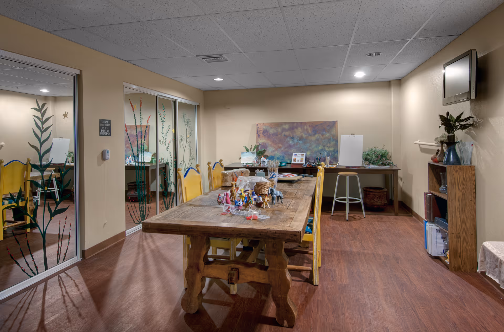 A well-lit room with a large wooden table surrounded by yellow chairs. The table has various art supplies and small sculptures on it. The room has wooden flooring, beige walls, and a ceiling with recessed lighting. There are glass sliding doors with painted plant designs on the left side, and a desk with art materials and a stool against the far wall. A small bookshelf with plants and books is on the right side, and a wall-mounted TV is above it.