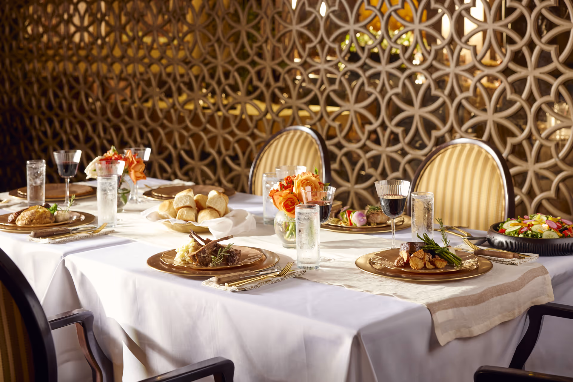 A dining table set with white tablecloth and beige runner, featuring plates with gourmet meals including lamb chops, steak with potatoes and asparagus, bread rolls, and a fresh salad. There are glasses of water and red wine, along with small vases of orange roses. The background has an intricate decorative wooden panel and upholstered chairs with striped beige fabric.