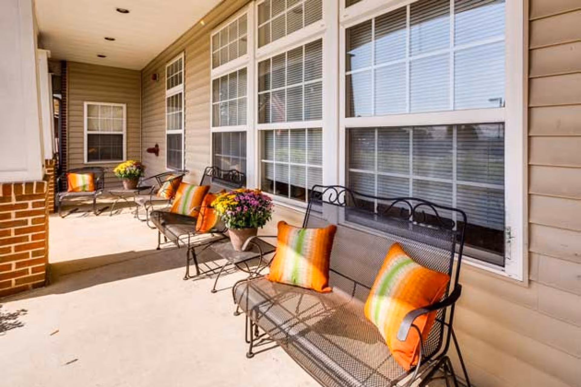 A covered outdoor patio area with metal benches and chairs adorned with orange and green striped cushions. There are potted flowers on small tables between the benches. The patio is adjacent to a building with beige siding and large windows with white frames and blinds.