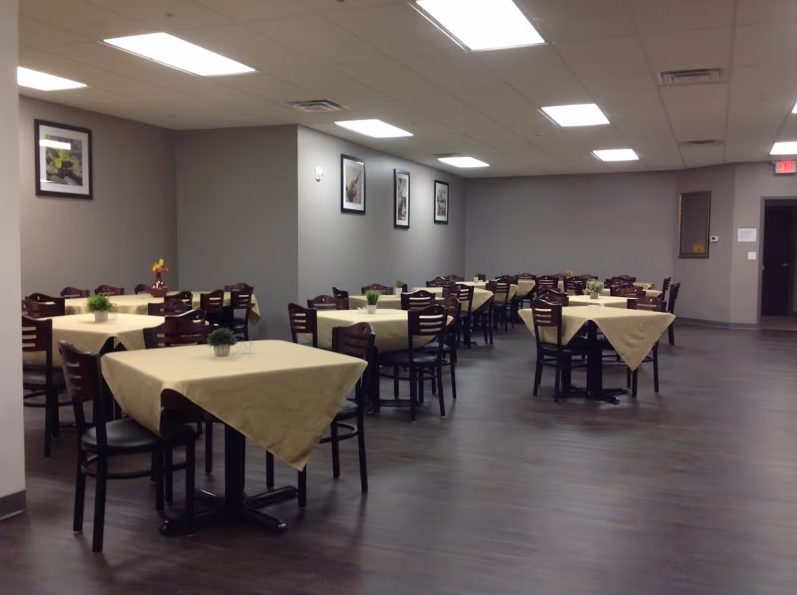 A dining room in Hyde Park Assisted Living Facility with multiple square tables covered with beige tablecloths. Each table is surrounded by dark wooden chairs and decorated with small green plants or floral arrangements. The room has gray walls adorned with framed pictures and a dark wood floor. The ceiling has recessed lighting panels.