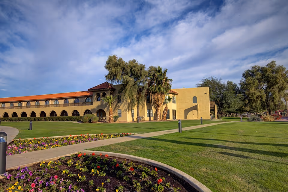 Exterior view of The Garnet of Casa Grande building with a yellow facade, red-tiled roof, and arched windows. The foreground features a well-maintained lawn, flower beds with colorful flowers, palm trees, and a partly cloudy sky.