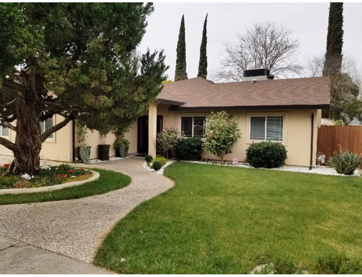 Front exterior view of a single-story beige house with a brown shingle roof, surrounded by green grass, shrubs, and trees. A curved concrete pathway leads to the front door, and there are two tall cypress trees behind the house.