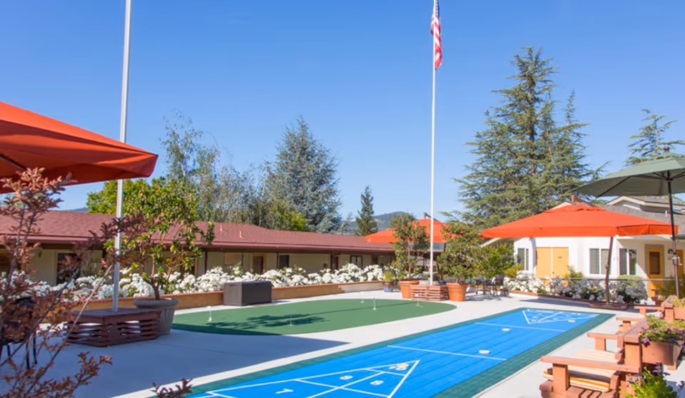 Outdoor recreational area at a senior living facility with a shuffleboard court, putting green, large red umbrellas, potted plants, benches, and an American flag on a flagpole under a clear blue sky.