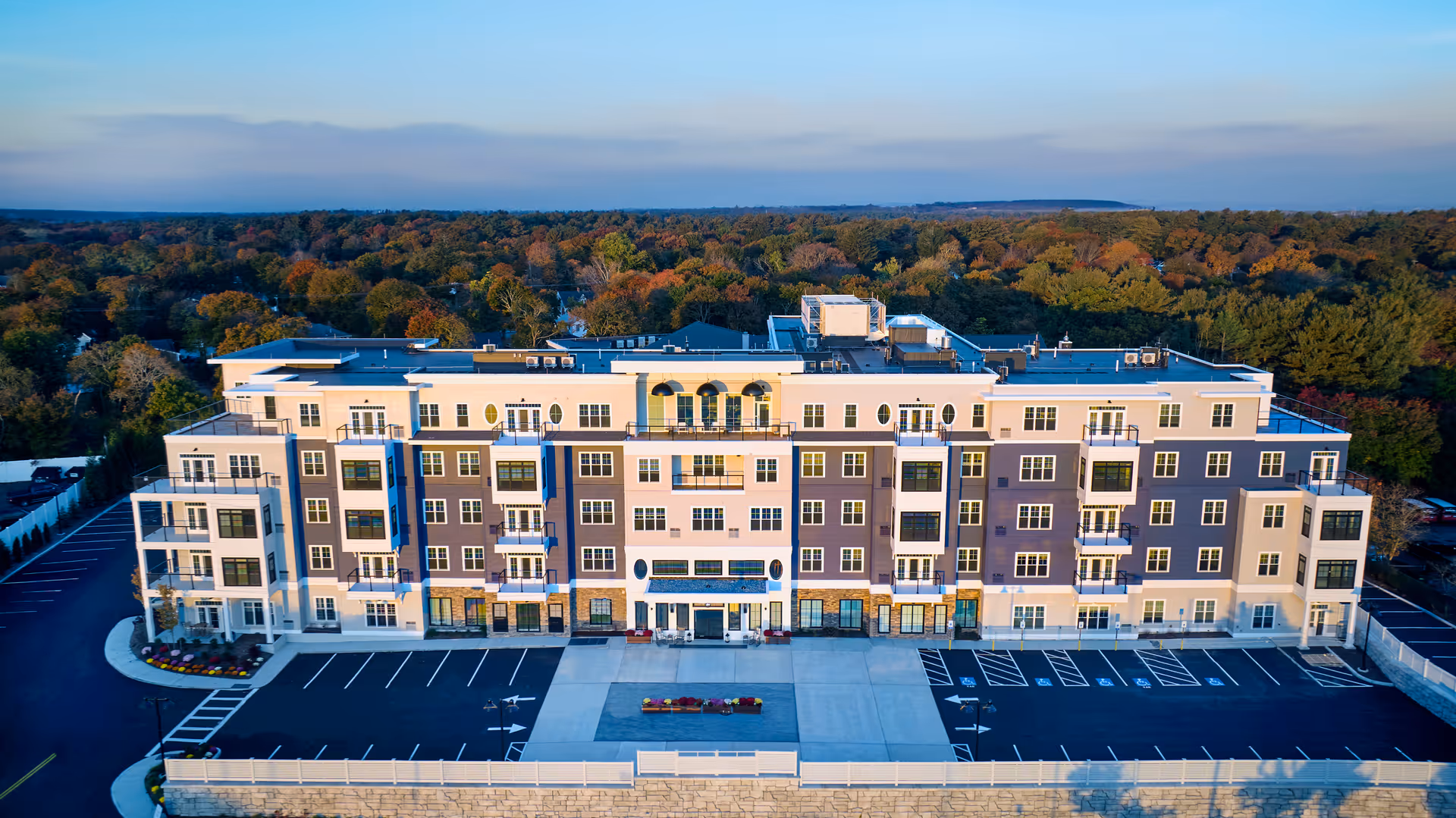 Aerial front view of a large multi-story senior living building with a paved parking lot and treeline in the background.