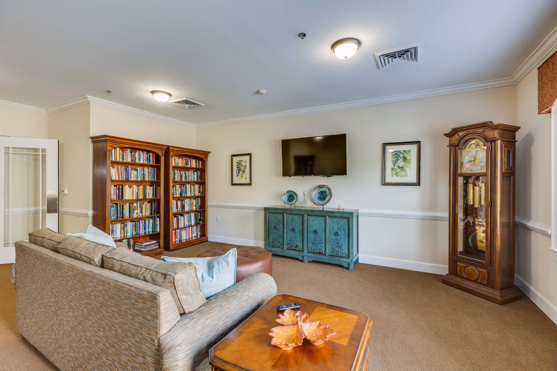 A cozy living room with a patterned sofa, a wooden coffee table with decorative bowls, a large wooden bookshelf filled with books, a turquoise cabinet with decorative plates on top, a wall-mounted flat-screen TV, two framed botanical prints, and a tall wooden grandfather clock near a window.