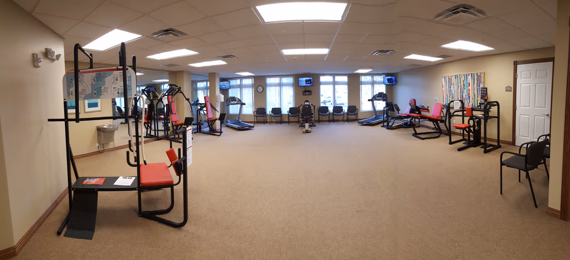 A spacious fitness room in Cahaba Ridge Retirement Community with various exercise equipment including treadmills, weight machines, and chairs arranged around the room. The room has beige walls, carpeted floor, and large windows letting in natural light. There is a colorful abstract painting on one wall and ceiling lights providing bright illumination.