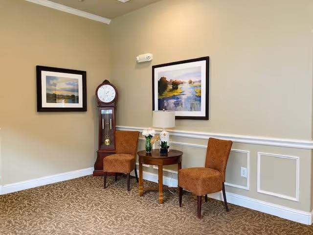 A cozy corner of a room with two brown upholstered chairs positioned around a small wooden round table. On the table are a lamp, a small potted plant, and a vase with flowers. Behind the chairs, there is a tall grandfather clock and two framed landscape paintings hanging on beige walls with white trim. The floor is carpeted with a patterned design.