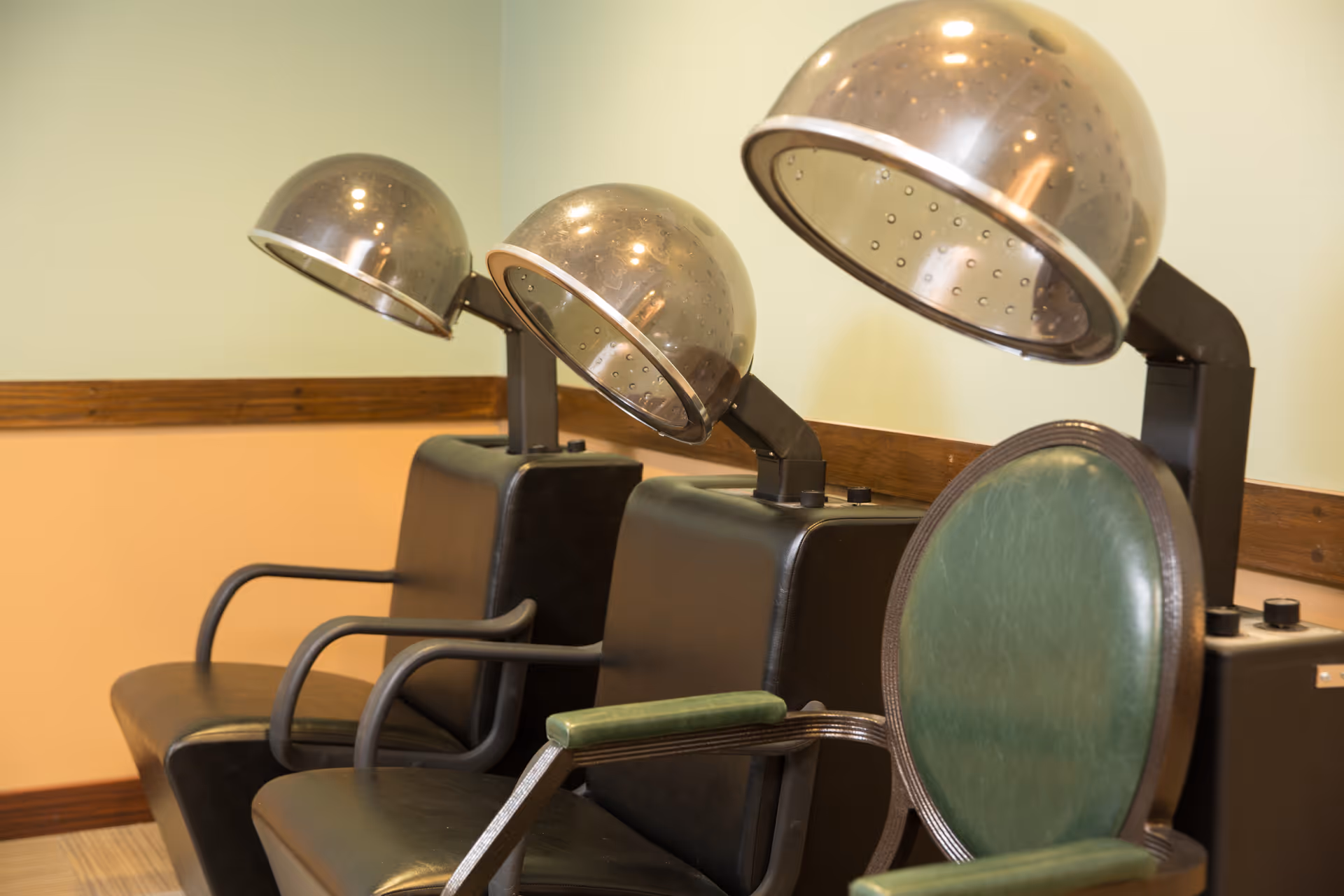 Three vintage hair drying chairs with large dome-shaped hair dryers in a salon area with light green and beige walls and wooden trim.