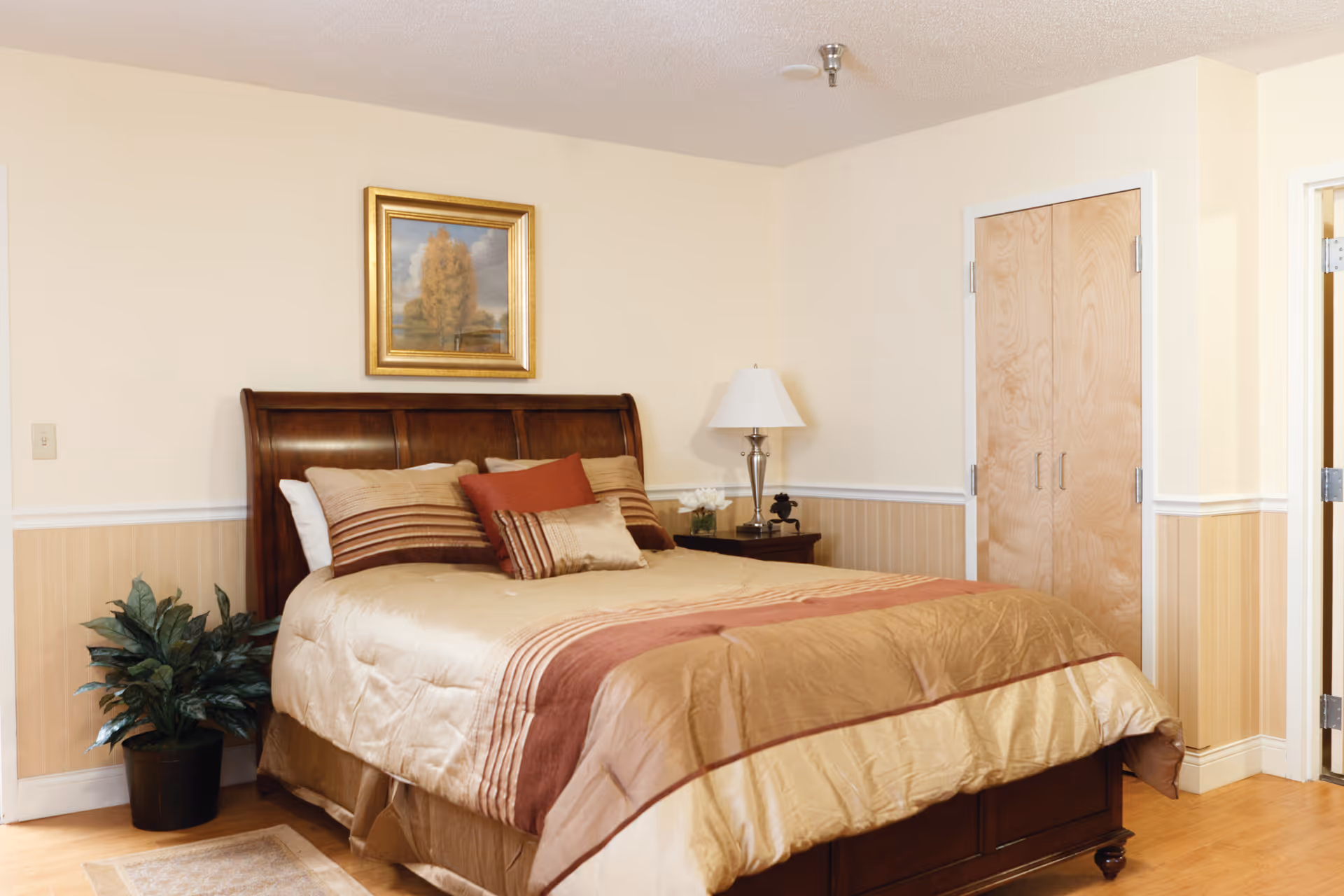 A neatly made bed with beige and brown bedding in a bedroom. The bed has multiple pillows and a wooden headboard. There is a nightstand with a lamp and decorative items to the right of the bed. A framed painting hangs on the wall above the bed. A potted plant is placed on the floor to the left of the bed. There is a closed wooden closet door and an open door leading to another room on the right side of the image.