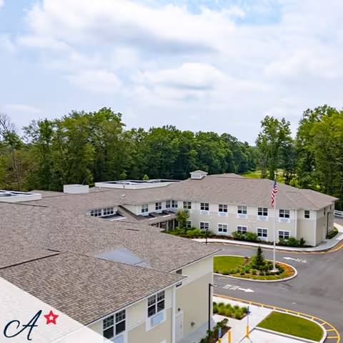 Aerial view of a large two-story assisted living facility building with beige exterior walls and a brown shingled roof, surrounded by greenery and trees under a partly cloudy sky. There is a circular driveway with an American flag on a flagpole near the entrance.