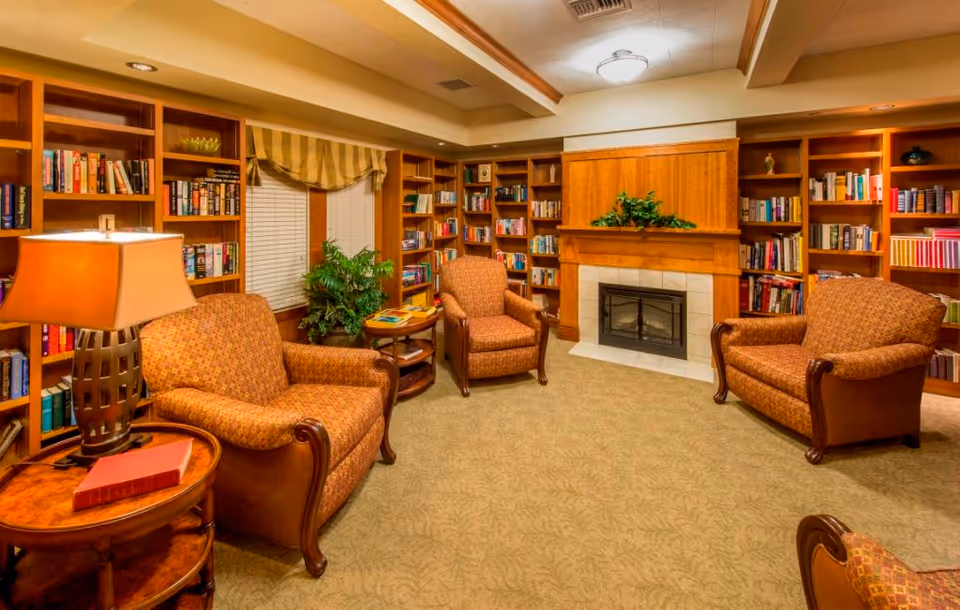 A cozy senior living community library or lounge area with three upholstered armchairs arranged around a fireplace. The room features built-in wooden bookshelves filled with books, a side table with a lamp and a book, a potted plant, and a window with striped curtains.