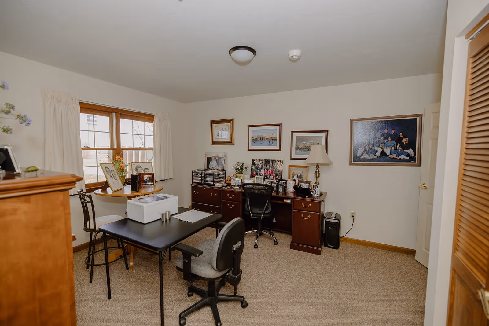 A cozy office room with beige walls and carpet. There is a large wooden desk with multiple framed photos, a lamp, and office supplies. In front of the desk is a black office chair. A smaller black table with two chairs is positioned near a window with cream-colored curtains. Several framed pictures and family photos hang on the walls.