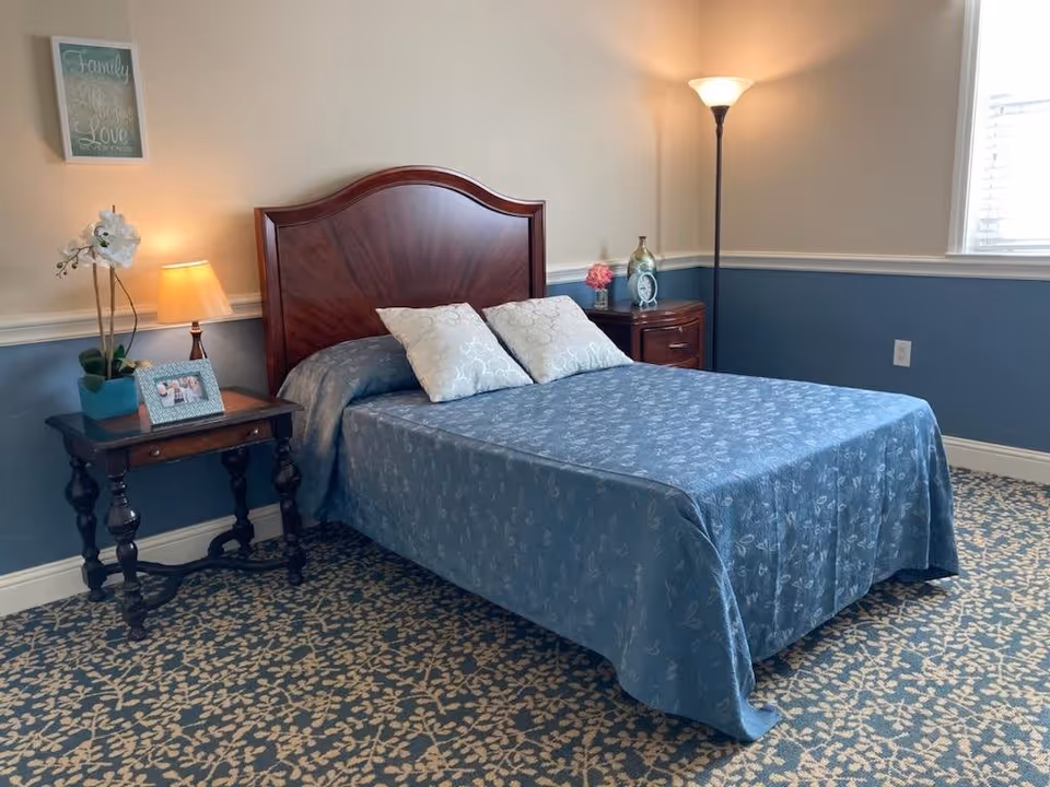 A neatly made bed with a wooden headboard in a bedroom. The bed has a blue patterned bedspread and two decorative pillows. On the left side of the bed is a small wooden side table with a lamp, a framed photo, and a potted white orchid. On the right side is a wooden nightstand with a clock, a small flower vase, and a tall floor lamp. The walls are painted beige on top and blue on the bottom with white trim, and there is a window with blinds letting in natural light. A framed sign on the wall reads 'Family, Life, Laughter, Love'. The floor is covered with a blue and beige patterned carpet.