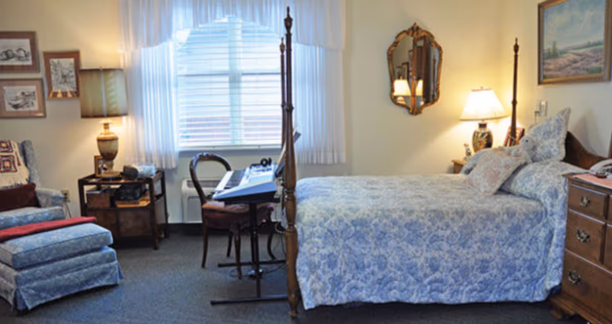 A cozy bedroom with a four-poster bed in blue patterned bedding, bedside tables and lamps, an armchair with ottoman, and a keyboard on a chair by the window.