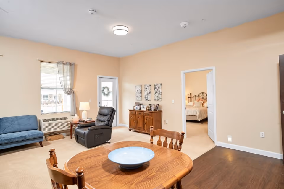 Bright furnished living and dining area with a wooden table and bowl, recliner, sofa, sideboard, and a doorway into a bedroom.