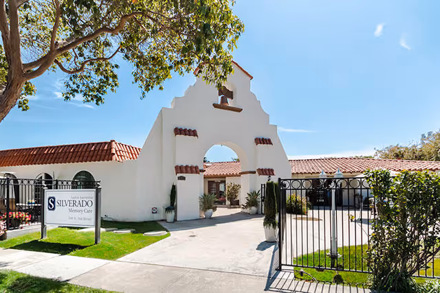 Exterior view of Silverado Tustin Hacienda Memory Care Community featuring a white stucco archway with red tile accents, a gated entrance, and a sign displaying the facility's name. The scene includes a tree, green lawn, and a clear blue sky.