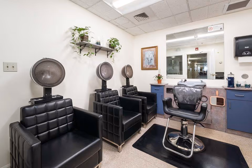 Interior of a salon area in a senior living facility with three black leather chairs under hair dryers, a black salon chair in front of a large mirror, blue cabinets, a small plant on the counter, and a shelf with potted plants on the wall.