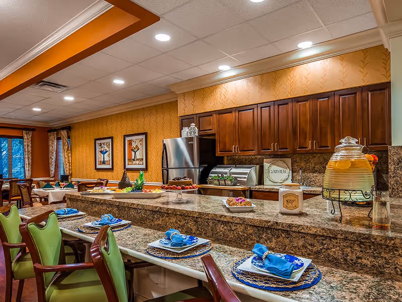 A dining area with a granite countertop bar set with plates, blue napkins, and green cushioned chairs. Behind the bar is a kitchen area with wooden cabinets, a stainless steel refrigerator, and various food items and decorations on the counter. The room has warm yellow wallpaper and framed artwork on the walls.