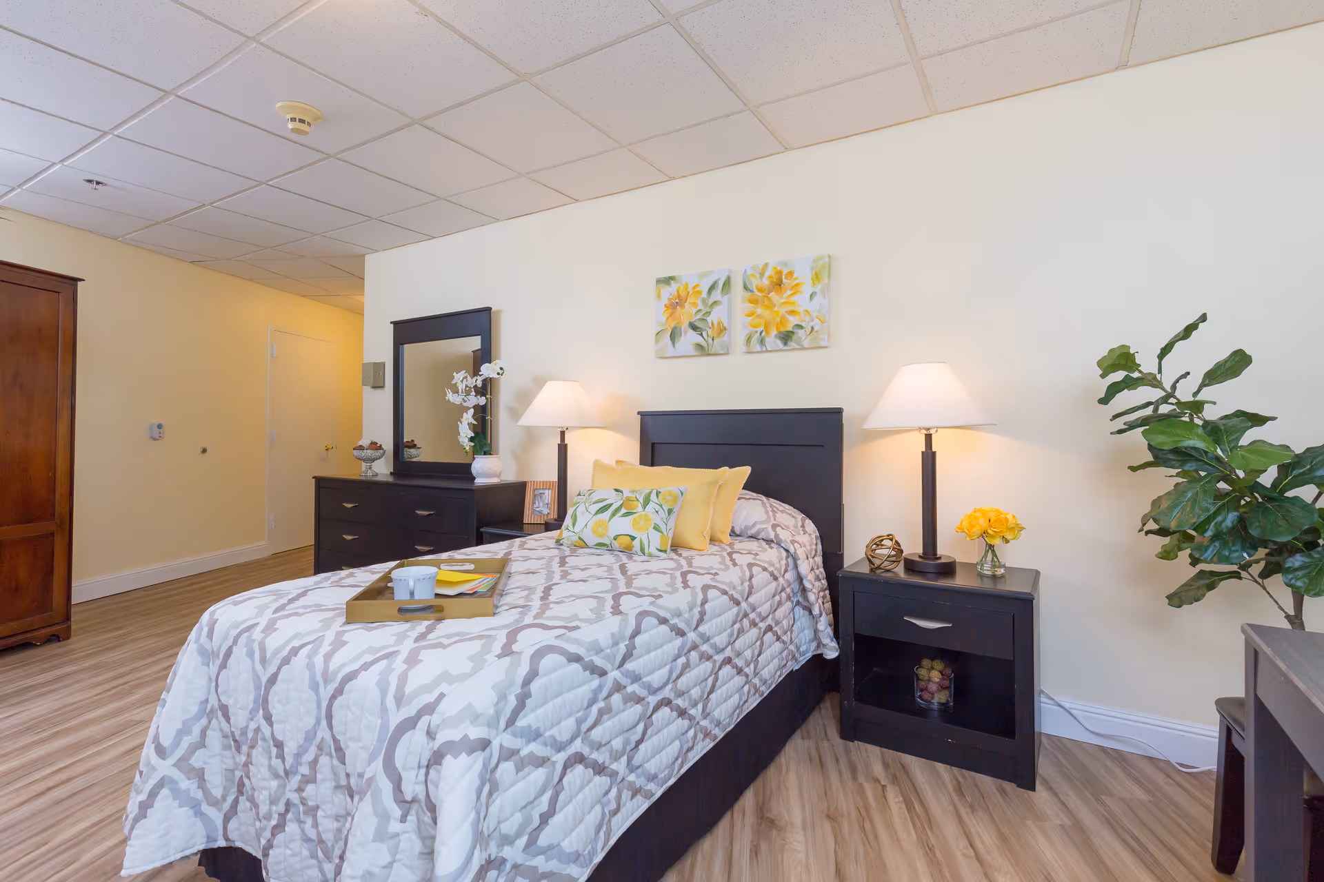 A neatly arranged bedroom in a senior living facility featuring a single bed with a patterned quilt and yellow accent pillows. On either side of the bed are black nightstands with lamps, decorative items, and a small vase of yellow flowers. A black dresser with a mirror and a white orchid plant is positioned against the wall. The room has light-colored walls, wood flooring, and two floral paintings above the bed. A green leafy plant is placed near the nightstand on the right.