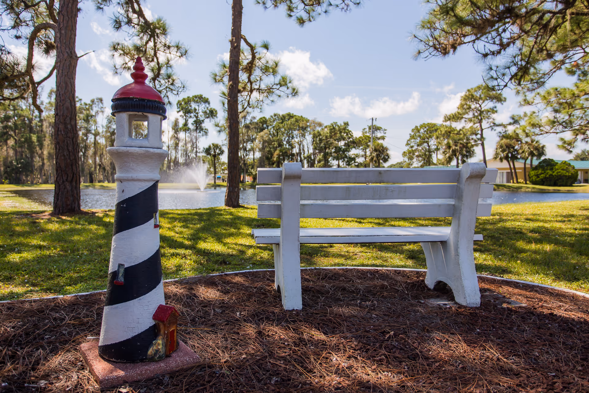 A white wooden bench next to a small decorative lighthouse statue in a grassy outdoor area with pine trees and a pond with a water fountain in the background under a partly cloudy sky.