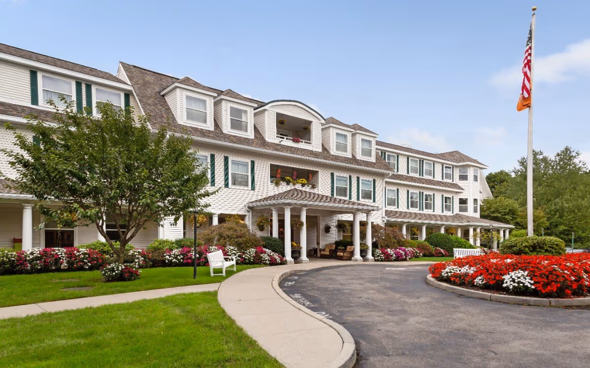 Exterior view of a large, white senior living facility building with multiple windows and a covered entrance. The building is surrounded by well-maintained landscaping including green grass, colorful flower beds, bushes, and trees. There is a curved driveway leading to the entrance and an American flag on a flagpole near the flower bed.